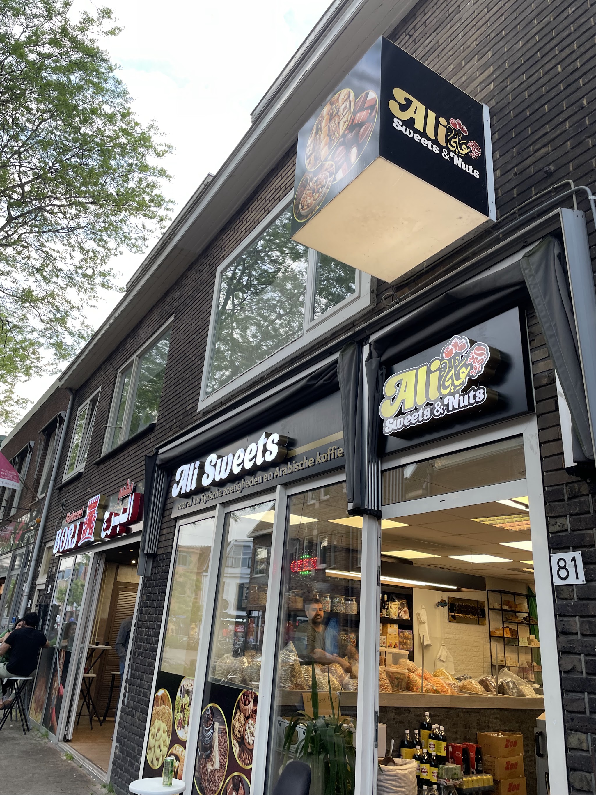 Gevel van Ali Sweets & Nuts met de uithangbord, interieur zichtbaar met medewerker achter de toonbank